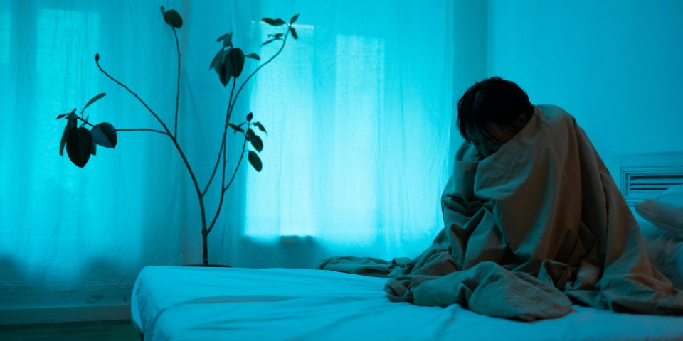 A woman sits alone on a bed wrapped in a blanket, conveying loneliness and depression.