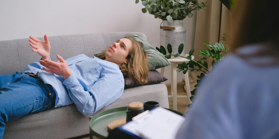 Concerned male client lying on sofa and speaking about mental problems with unrecognizable psychologist with clipboard during psychotherapy appointment in office