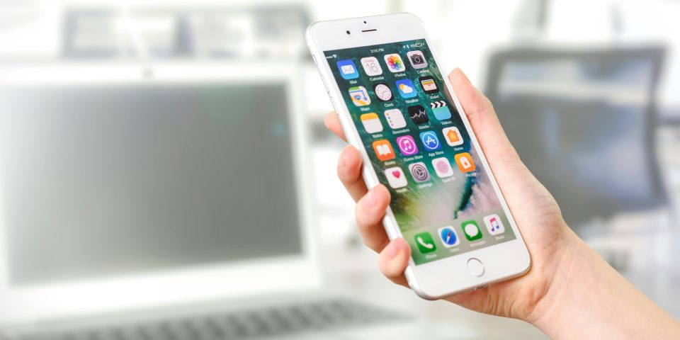 Close-up view of a smartphone displaying apps, held by a hand, with a blurred laptop in the background.